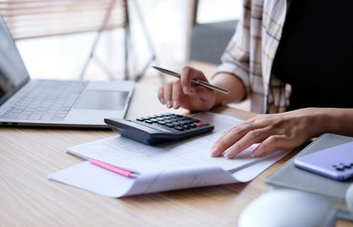 Closeup of the hands of a casually-dressed woman sitting at a desk covered with papers and tools related to personal finance