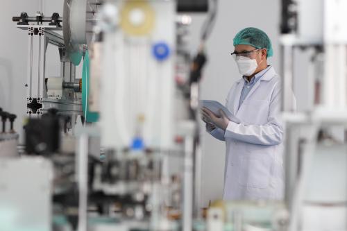 Worker man in personal protective equipment or PPE inspecting quality of mask and medical face mask production line in factory, manufacturing industry and factory concept.