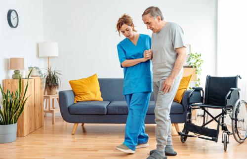 Young caregiver assisting senior man in wheelchair walking indoors - Home health care service and physiotherapy concept